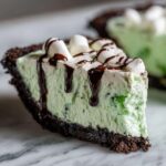 A close-up slice of creamy, mint-green grasshopper pie with a dark chocolate crust, topped with whipped cream, marshmallows, and chocolate syrup.