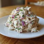 A mound of creamy greek yogurt chicken salad mixed with visible chunks of red onion and green herbs, served on a white plate.