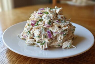 A mound of creamy greek yogurt chicken salad mixed with visible chunks of red onion and green herbs, served on a white plate.