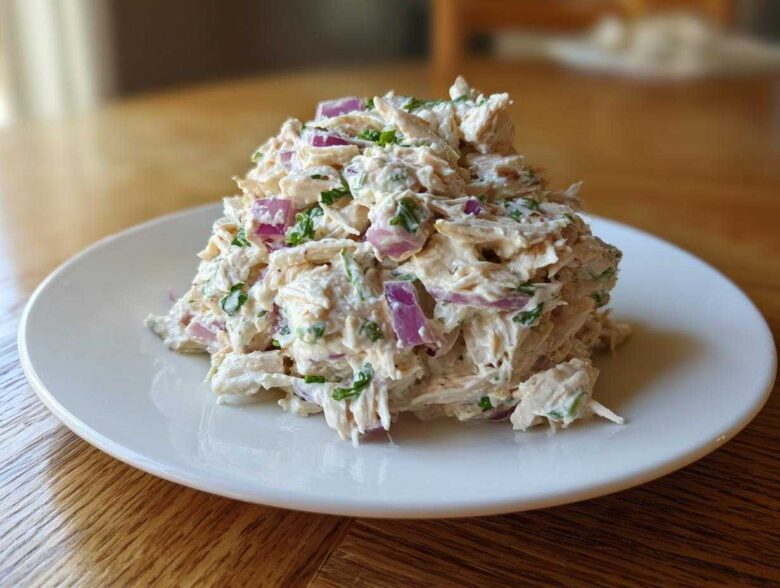 A mound of creamy greek yogurt chicken salad mixed with visible chunks of red onion and green herbs, served on a white plate.
