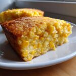 Close-up of a moist, golden-brown slice of jiffy corn casserole loaded with whole corn kernels on a white plate.