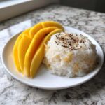 A white plate featuring sliced fresh mango next to a mound of sweet mango sticky rice topped with toasted sesame seeds.