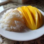 A plate of sweet mango sticky rice, featuring bright yellow sliced mango next to white sticky rice topped with sesame seeds.
