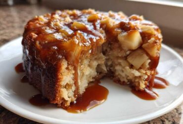 A close-up of a slice taken from a moist apple dapple cake, generously covered in a rich caramel sauce and visible apple chunks.