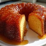 A moist Bacardi Rum Cake, glazed with a shiny caramel sauce, with one slice cut out on a white plate.