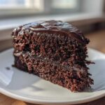 A close-up of a perfectly moist chocolate cake slice with glossy dark frosting on a white plate.