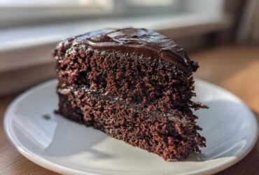A close-up of a perfectly moist chocolate cake slice with glossy dark frosting on a white plate.