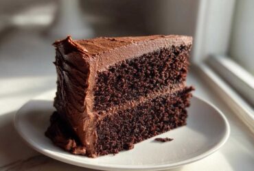 A decadent slice of moist chocolate cake layered with rich chocolate frosting, sitting on a white plate.