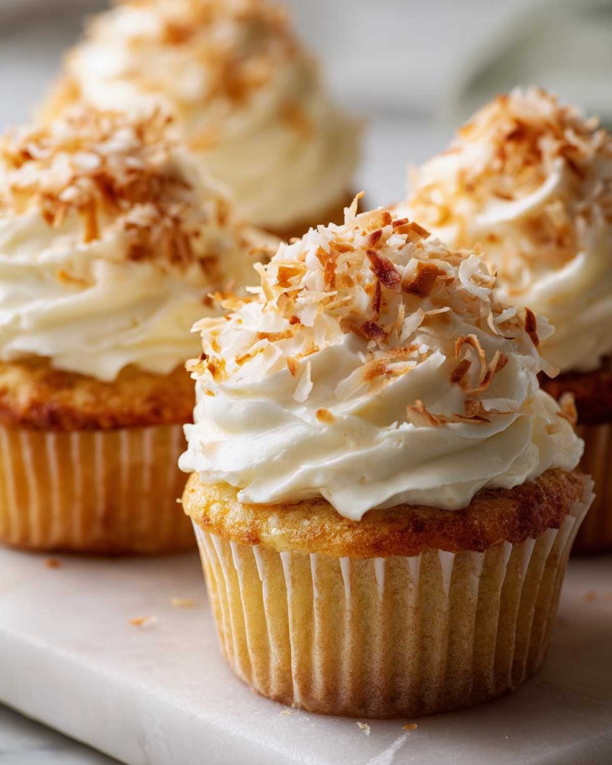 Close-up of moist coconut cupcakes topped with creamy white frosting and toasted coconut flakes.