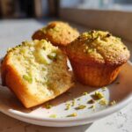 Close-up of moist pistachio muffins, one cut open to show the texture, topped with chopped pistachios.