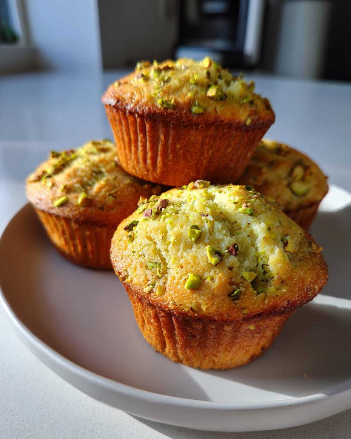 Four golden brown pistachio muffins stacked slightly on a white plate, topped with chopped green pistachios.