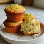 Close-up of three moist pistachio muffins, one cut open to show the crumb texture with green pistachio pieces.
