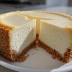A whole, crack-free new york cheesecake with two slices cut out, showing a thick graham cracker crust.