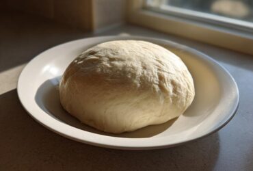 A smooth, pale ball of prepared fufu dough resting on a white plate, illuminated by sunlight near a window.
