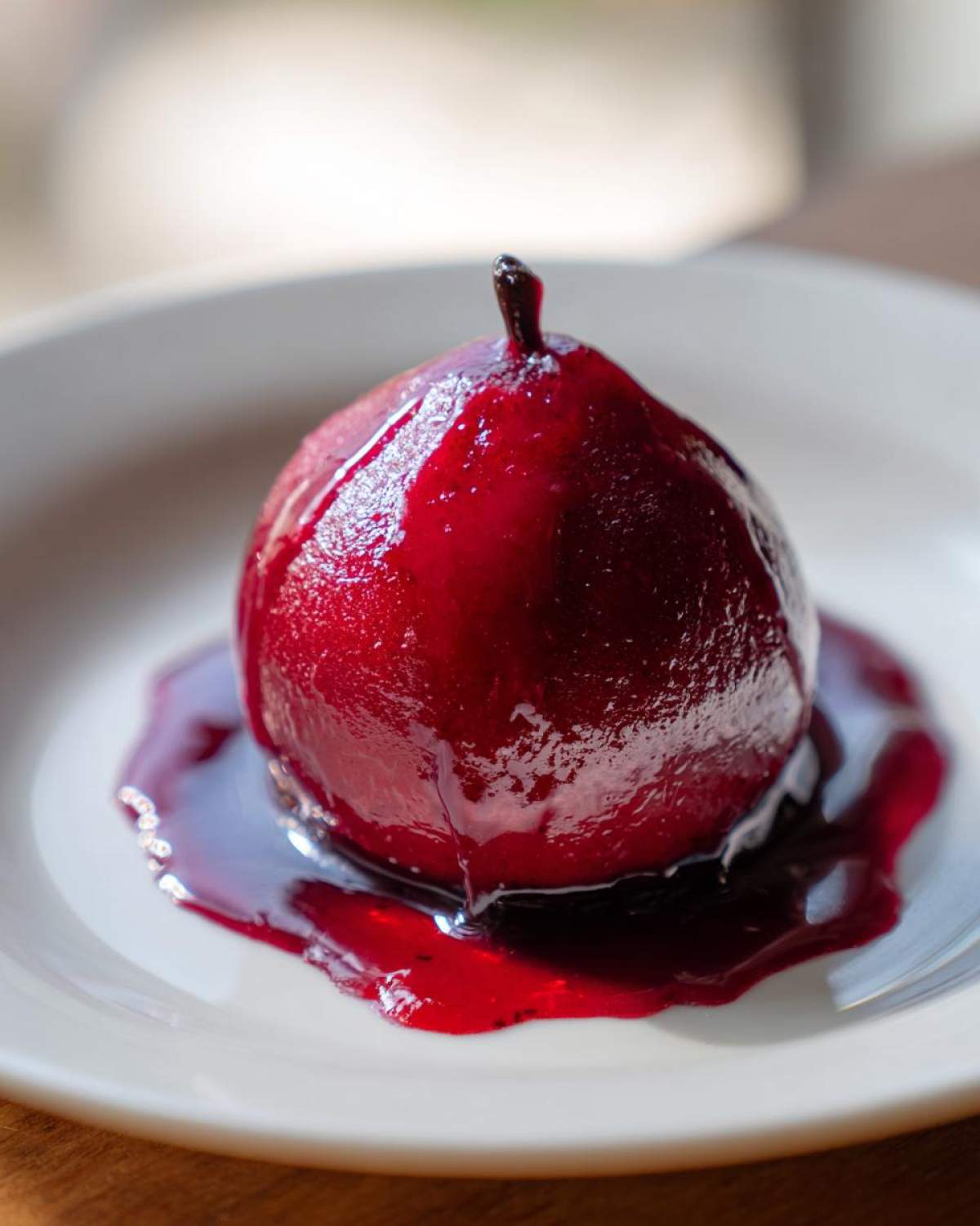 A single, glossy, deep red wine poached pears dessert served on a white plate with excess syrup.