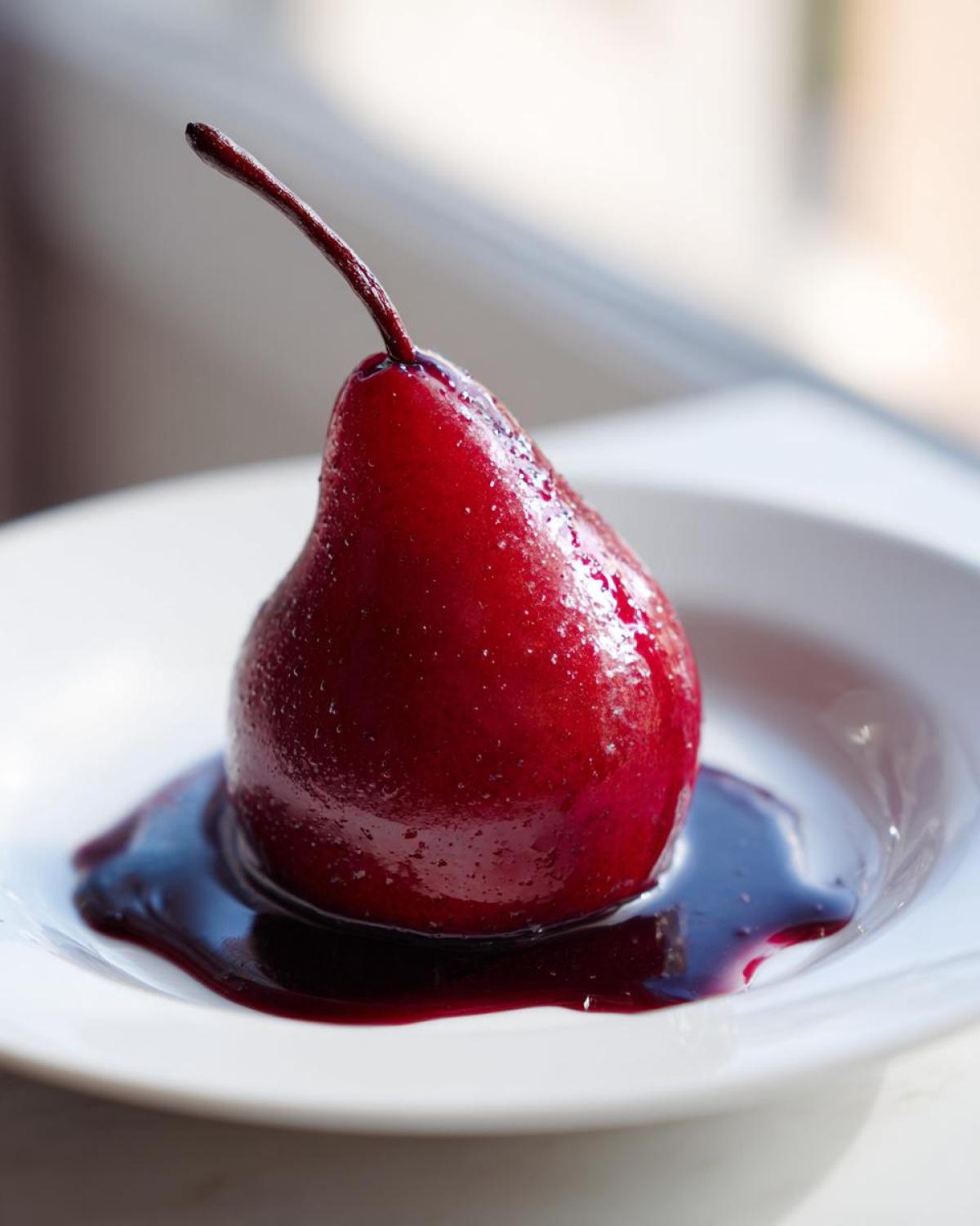 A single, vibrant red wine poached pears dessert sitting in a pool of deep red syrup on a white plate.