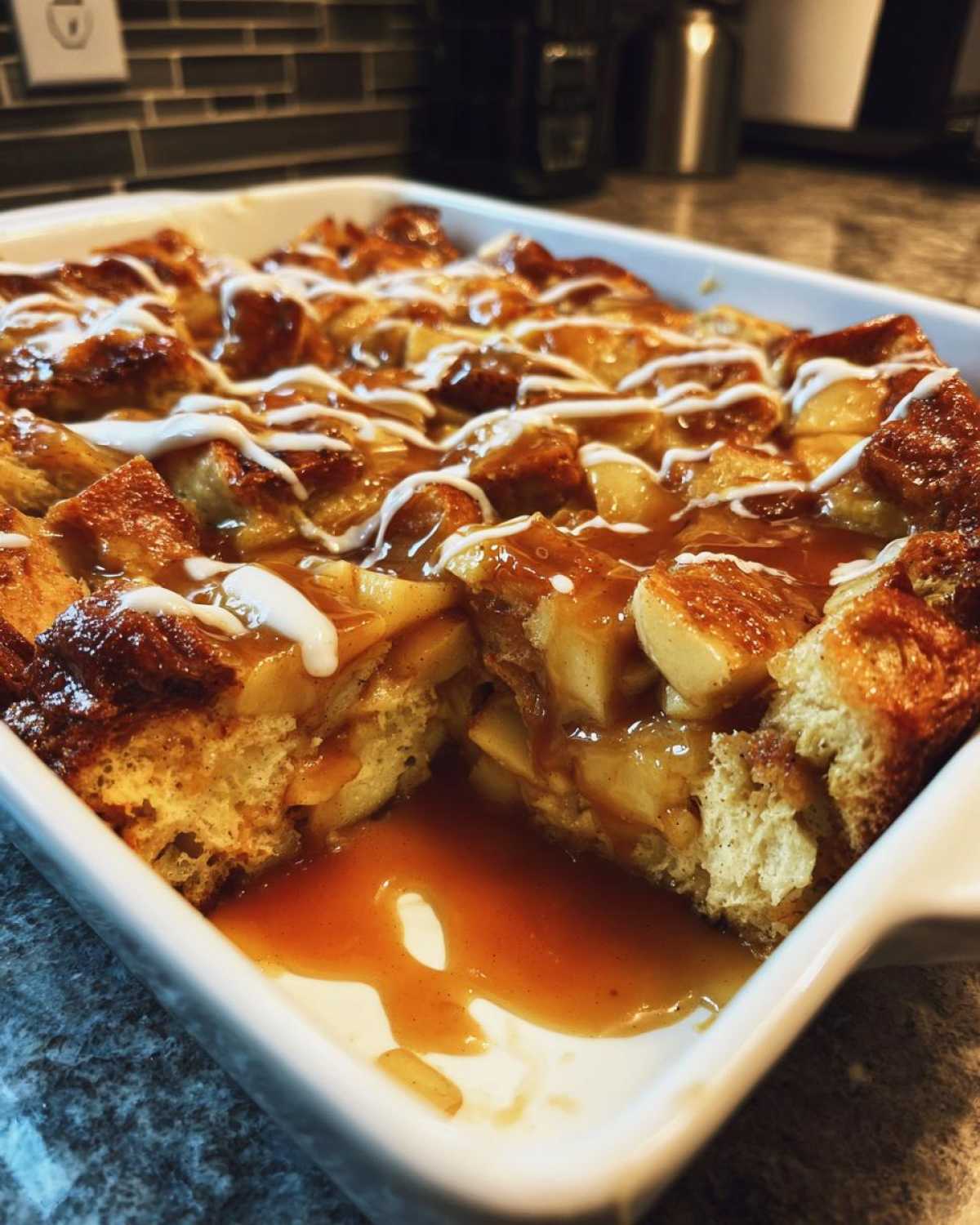 A square white baking dish filled with warm apple bread pudding, drizzled with caramel and white icing.
