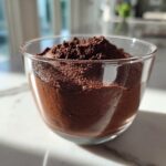 A close-up of rich, dark chocolate mousse topped with cocoa powder in a clear glass serving cup.