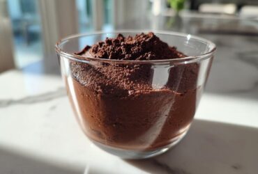 A close-up of rich, dark chocolate mousse topped with cocoa powder in a clear glass serving cup.