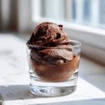 A scoop of rich, dark chocolate mousse served in a small clear glass, brightly lit by natural light.