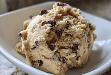 Close-up of a large scoop of creamy, light brown high protein cookie dough loaded with mini chocolate chips, served in a white bowl.