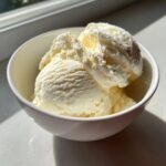 Three scoops of creamy, pale yellow cottage cheese ice cream served in a white bowl near a window.
