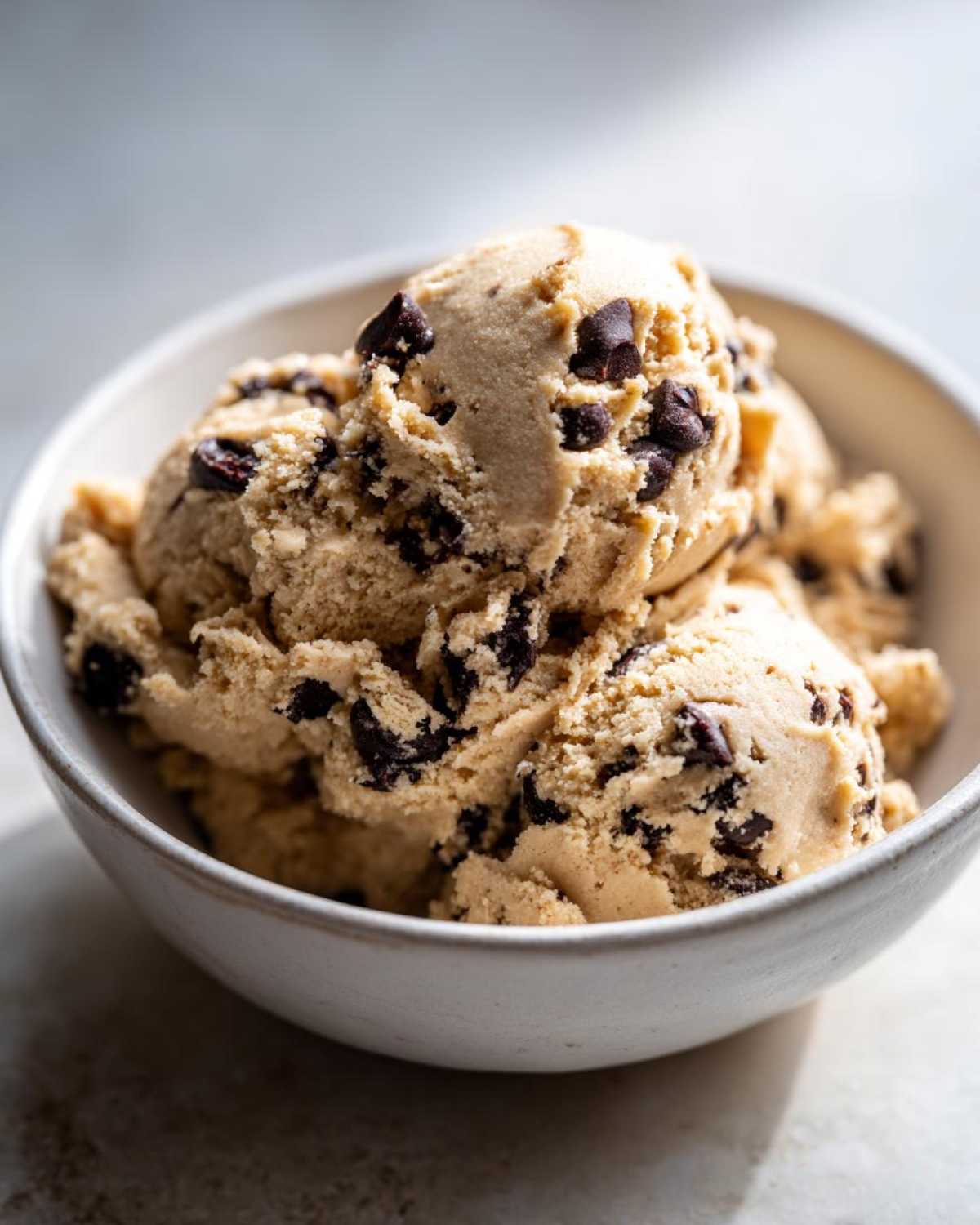 Three scoops of thick, light brown high protein cookie dough studded with dark chocolate chips served in a white bowl.