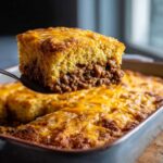 A spatula lifts a thick, layered slice of tamale pie, showing the cornbread top and savory meat filling.