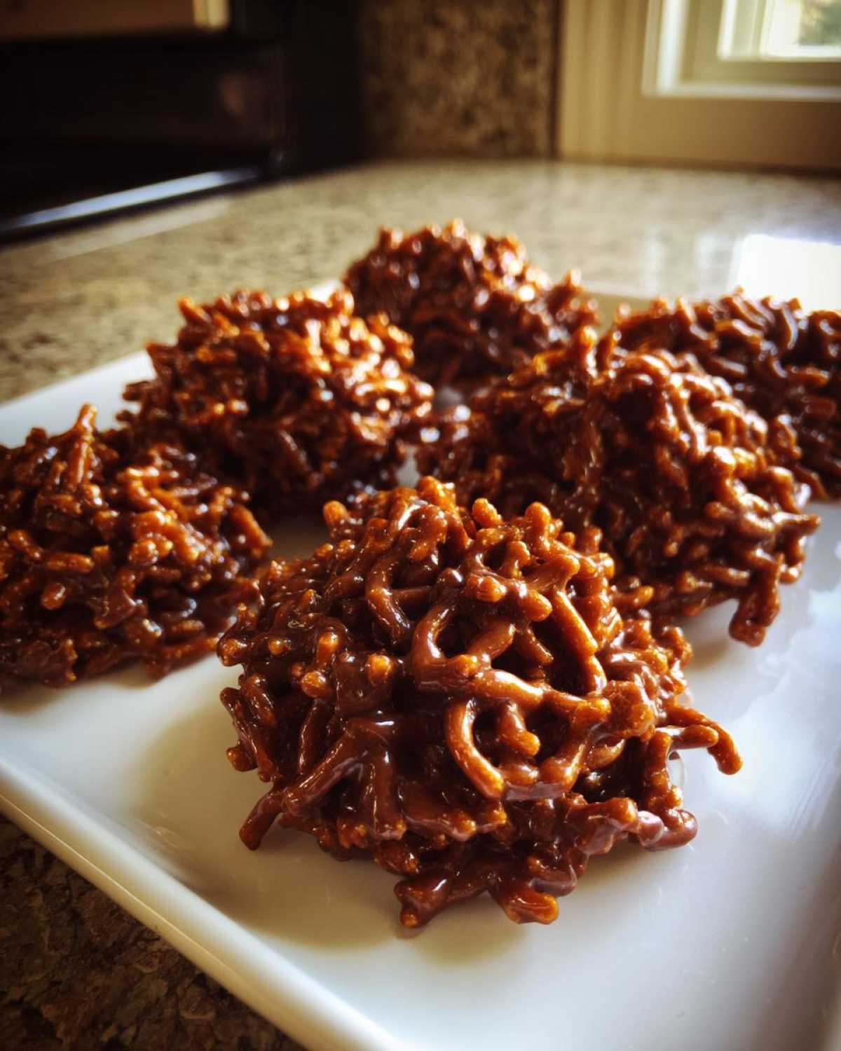 A close-up shot of several glossy, chocolate haystack cookies piled on a white rectangular plate.