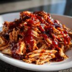 Close-up of shredded bbq chicken generously topped with rich, dark barbecue sauce on a white plate.