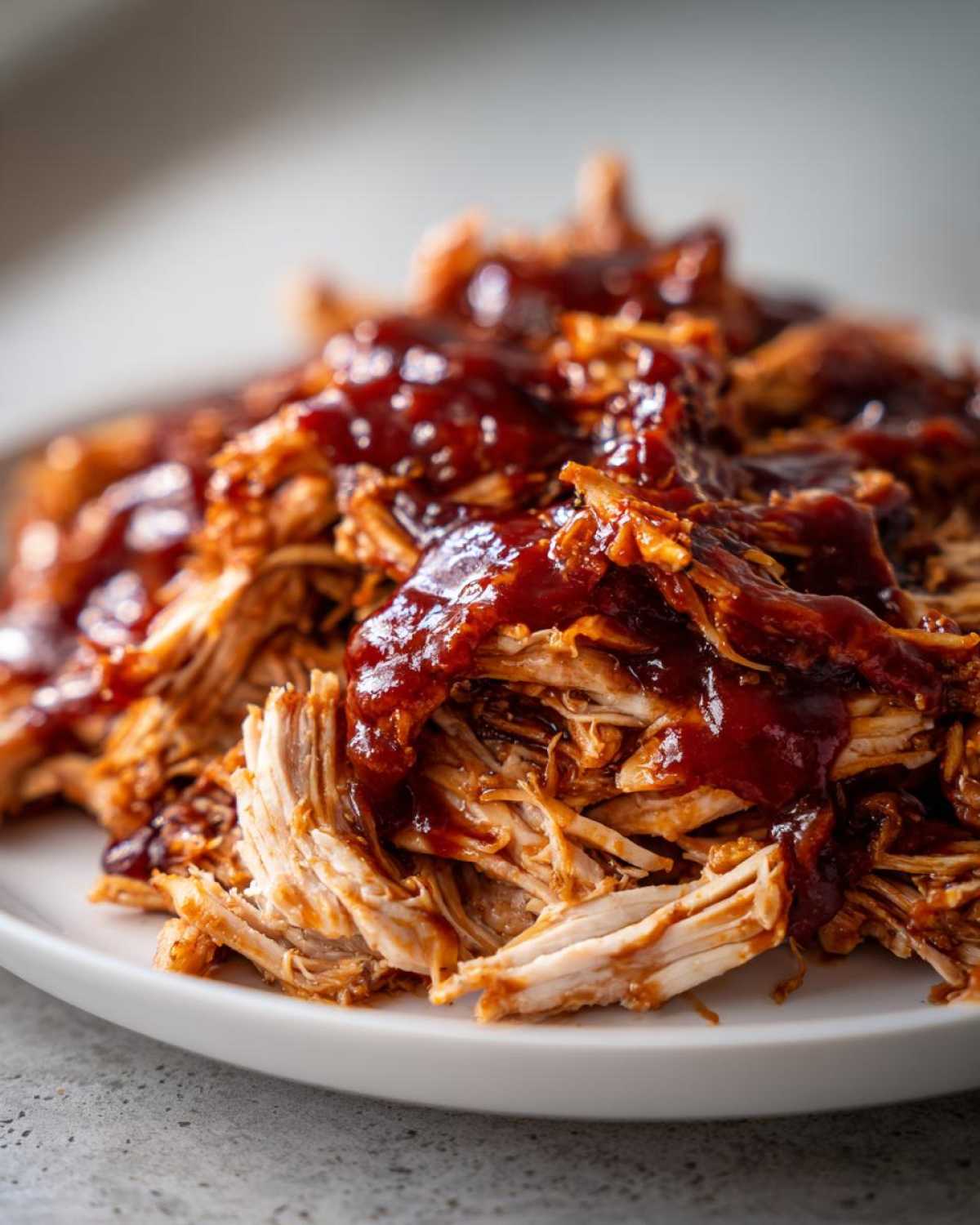 Close-up of tender shredded bbq chicken generously topped with rich, dark barbecue sauce on a white plate.