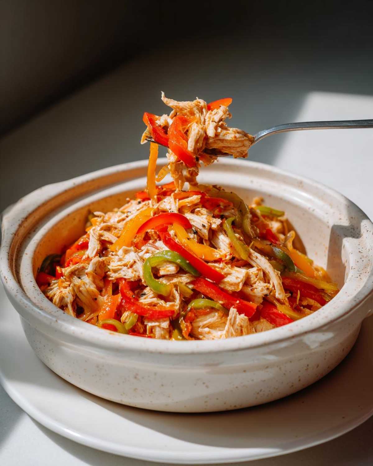 A fork lifts shredded chicken mixed with colorful bell pepper strips from a bowl of crockpot chicken fajitas.