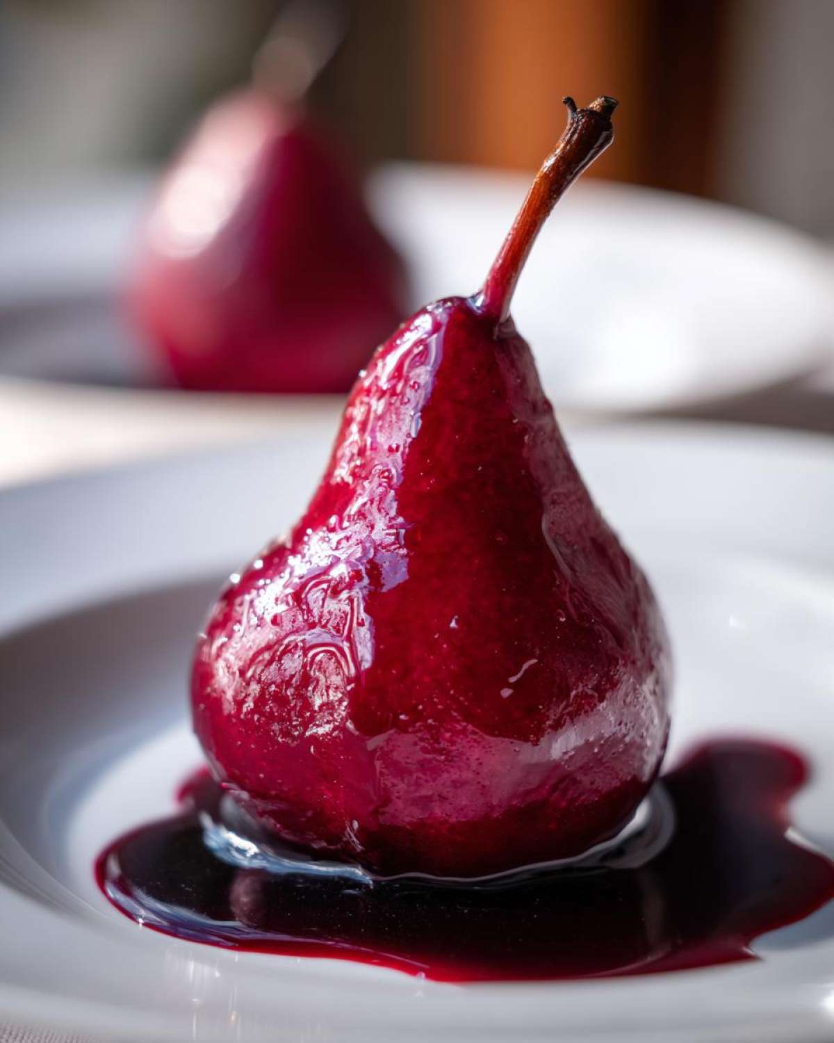 A glistening, deep red poached pear served on a white plate with rich syrup pooling around the base.