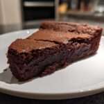 A dense, dark slice of flourless chocolate cake resting on a white plate, showing its fudgy texture.