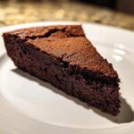 A close-up of a dark, rich slice of flourless chocolate cake dusted with cocoa powder, served on a white plate.
