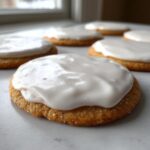 A close-up of a soft, golden cookie topped with a generous layer of thick, glossy white cookie icing.