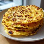 A stack of four golden yellow, freshly cooked soft corn tortillas with charred spots, resting on a light ceramic plate.