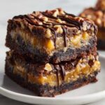 Two stacked, fudgy Turtle Brownies featuring a thick caramel layer, topped with pecans and a chocolate drizzle.