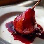 A single, whole poached pears dessert, deeply stained red by wine syrup, resting on a white plate with star anise.
