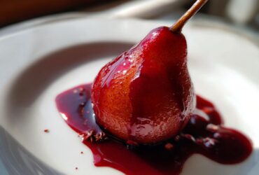 A single, whole poached pears dessert, deeply stained red by wine syrup, resting on a white plate with star anise.