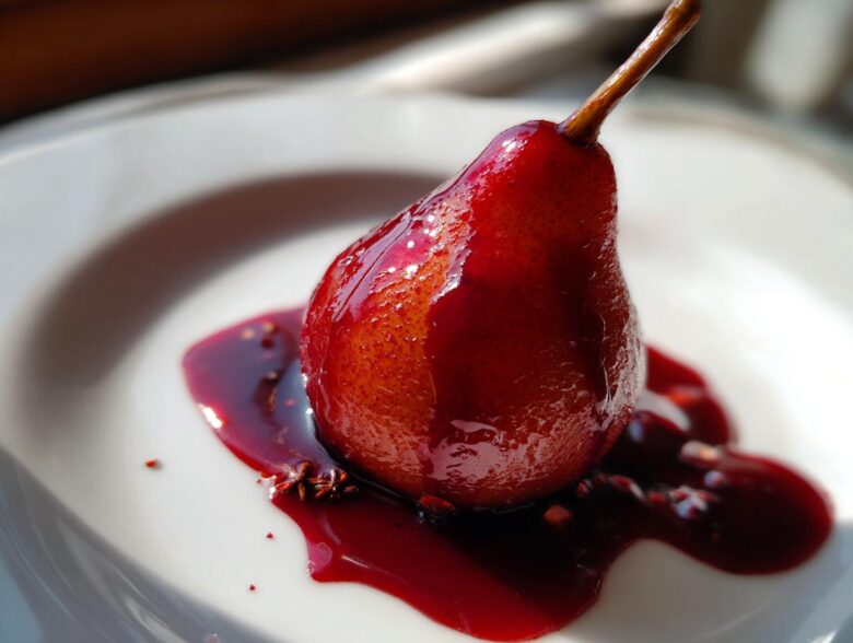 A single, whole poached pears dessert, deeply stained red by wine syrup, resting on a white plate with star anise.