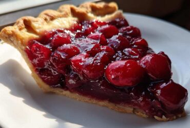 A close-up of a slice of pie generously topped with thick cherry pie filling and whole cherries.