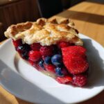 A close-up of a juicy slice of triple berry pie showing strawberries, blueberries, and raspberries, set against a flaky golden crust.