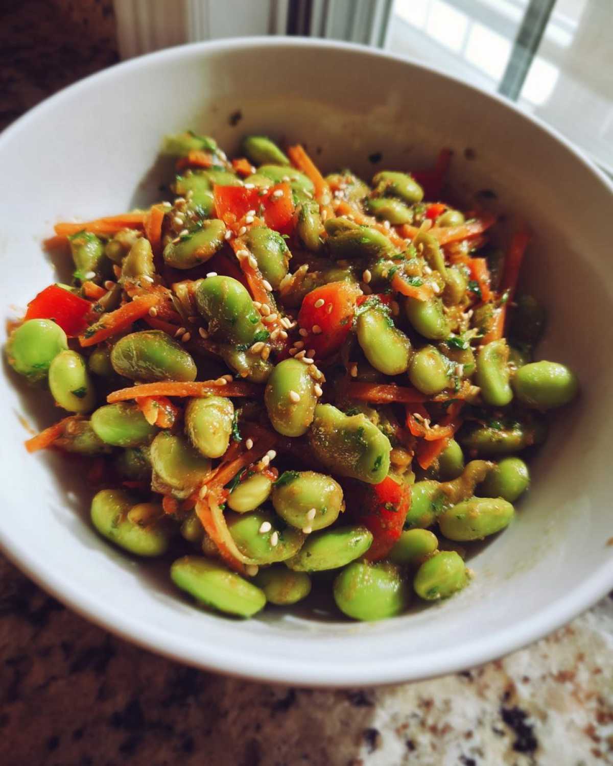 Close-up of a white bowl filled with a vibrant edamame salad mixed with shredded carrots, red peppers, and sesame seeds.