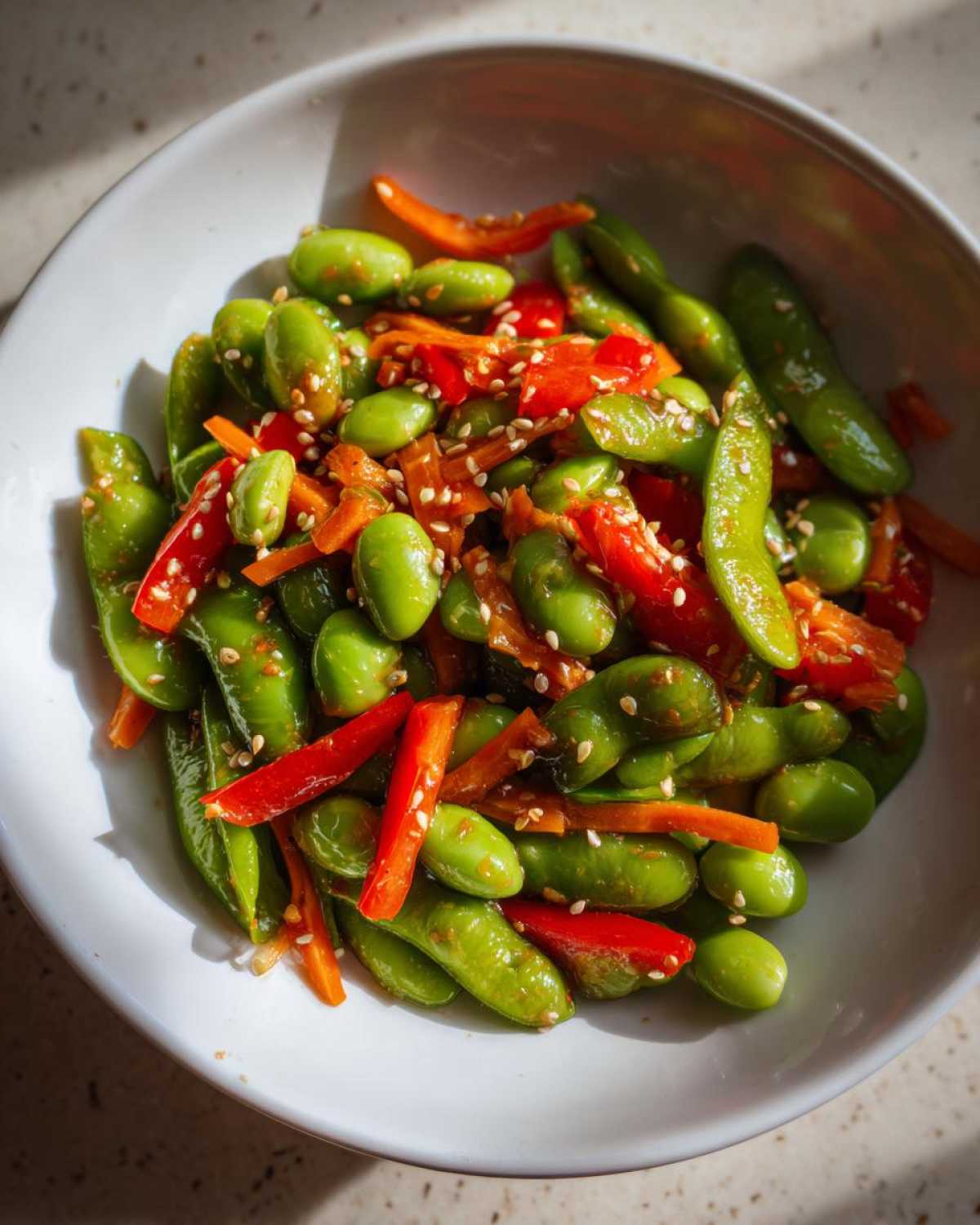 Close-up of a vibrant edamame salad featuring shelled edamame, bright red pepper strips, and sesame seeds in a white bowl.