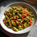 Close-up of a bright green edamame salad mixed with shredded carrots and sprinkled with sesame seeds in a white bowl.