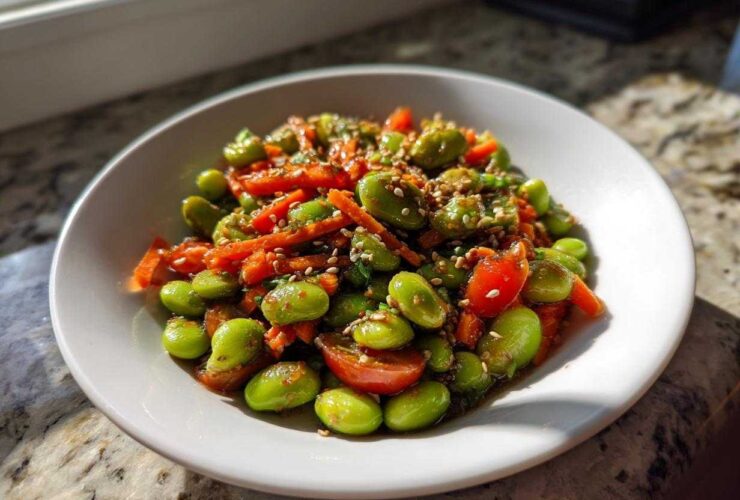 A close-up of a vibrant edamame salad with shredded carrots and halved cherry tomatoes, topped with sesame seeds.