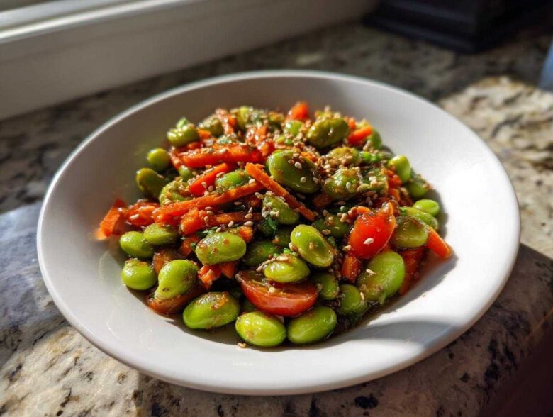 A close-up of a vibrant edamame salad with shredded carrots and halved cherry tomatoes, topped with sesame seeds.