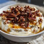A white bowl filled with creamy whipped goat cheese topped with candied pecans and drizzled with honey.