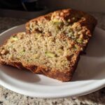 A close-up view of two slices of moist zucchini banana bread on a white plate, showing visible green zucchini pieces.